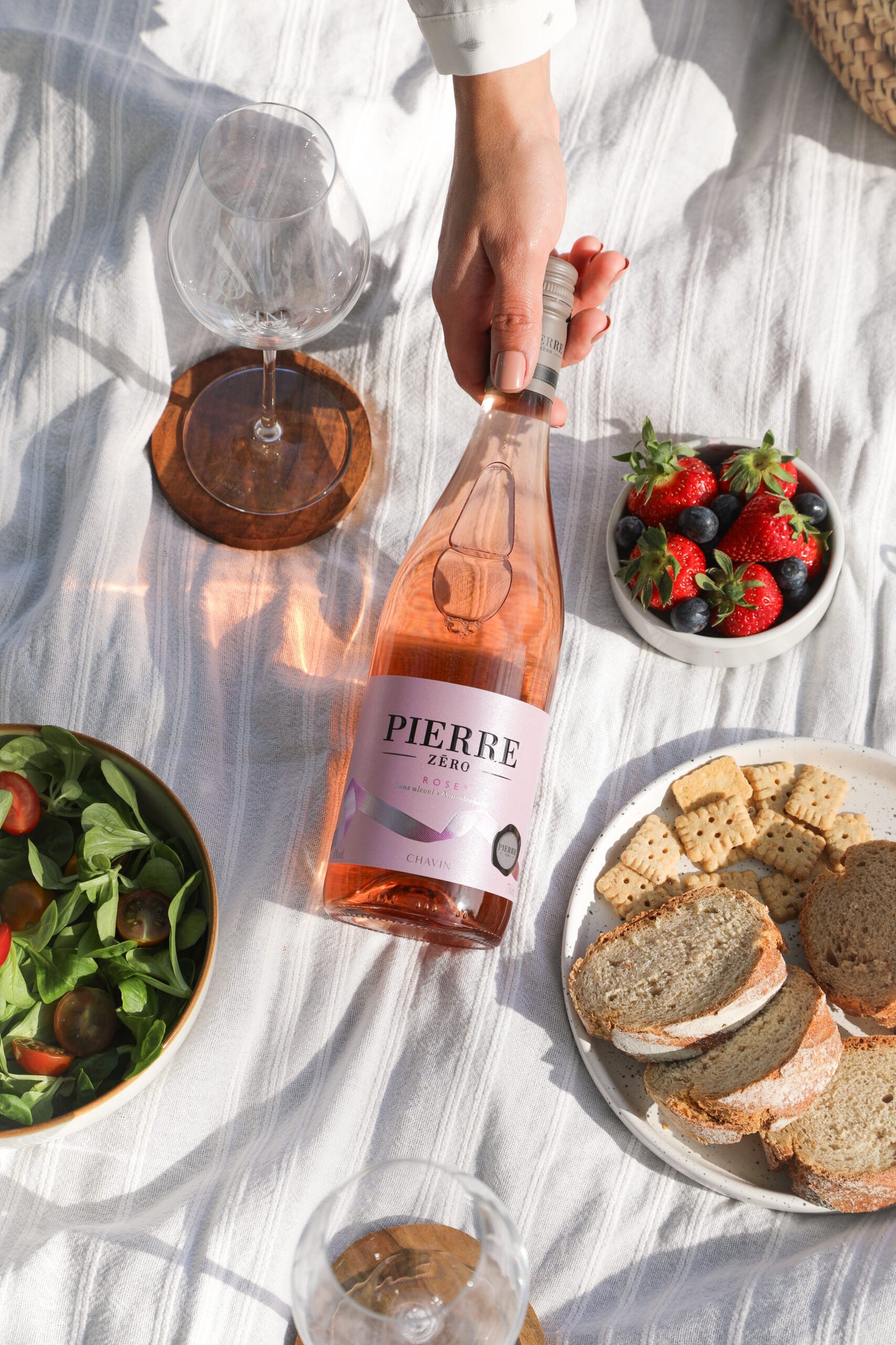 Person holding a bottle of 'Pierre' rosé wine with a picnic setup including bread, salad, and berries.