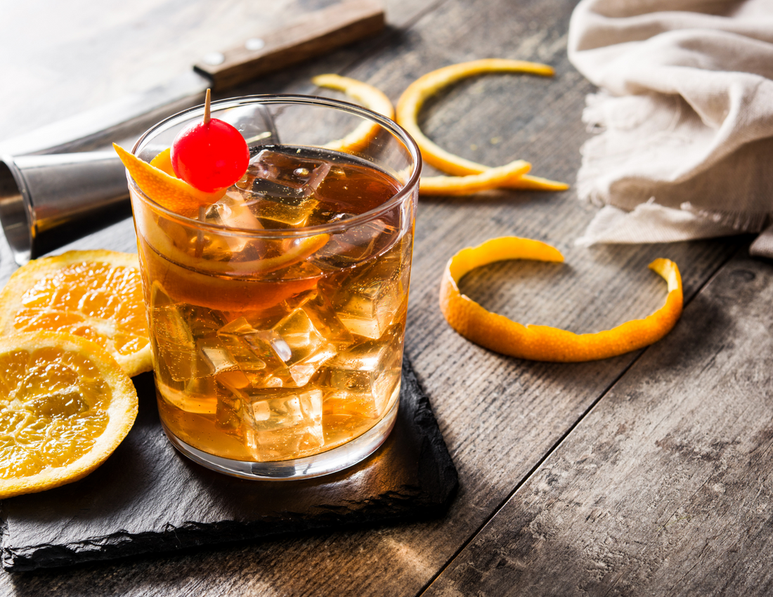Old Fashioned cocktail in a short glass with orange peel and cherry garnish. Orange slices and peels around. a jigger and knife in the background