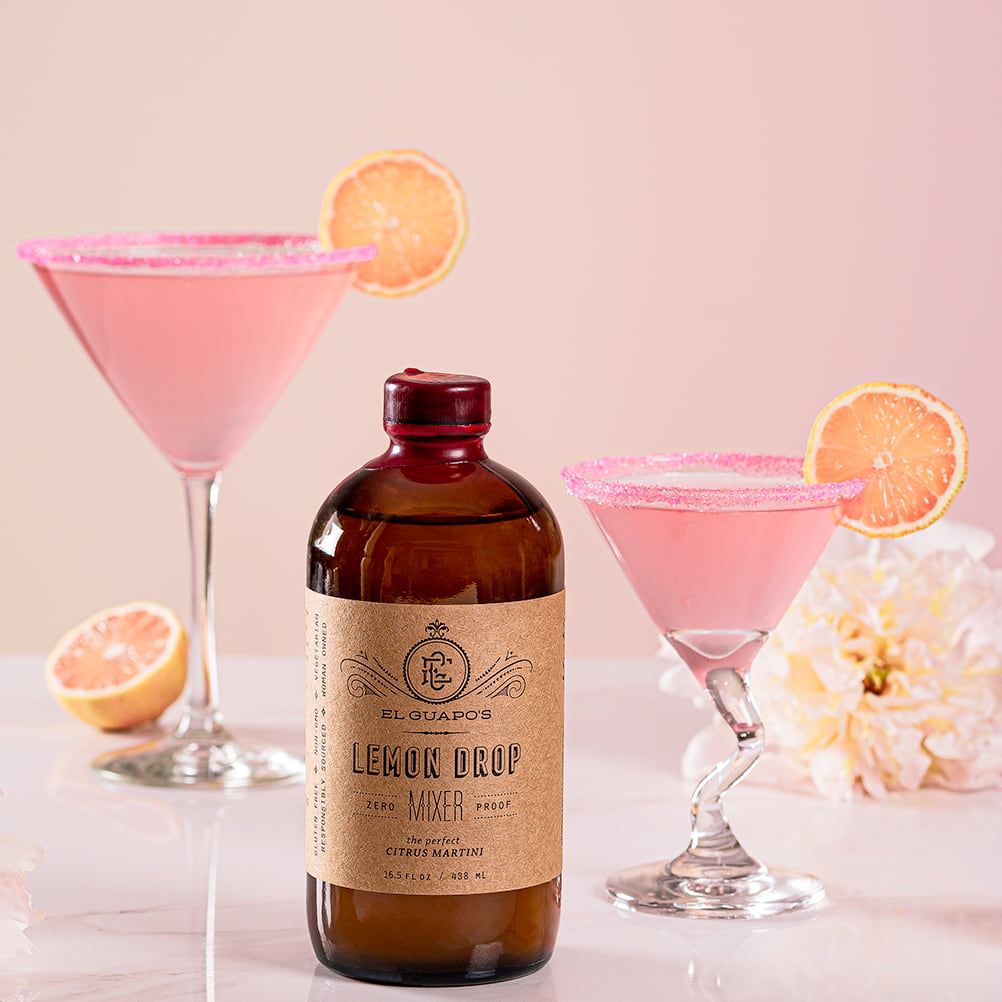 Bottle of El Chapo's Lemon Drop mixer with two pink cocktails and lemon slices on a light background